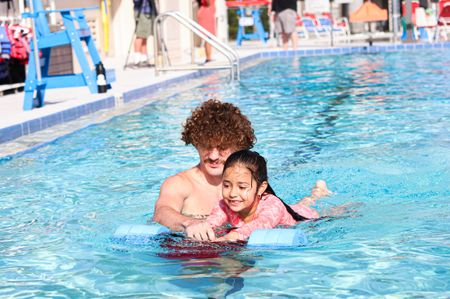 An instructor helping a little girl swim with a blue floaty during a CFY water safety program at a big outdoor pool . 