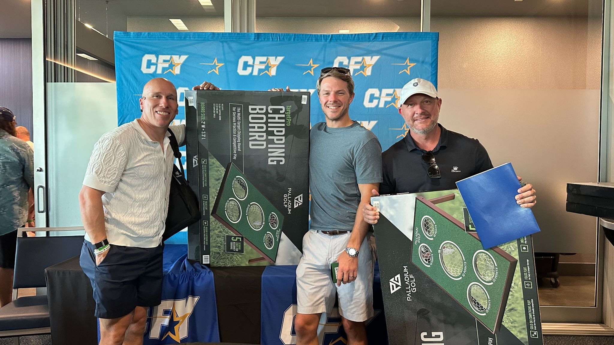 Three men holding golf game prizes at a CFY fundraising event in front of a CFY-branded backdrop.