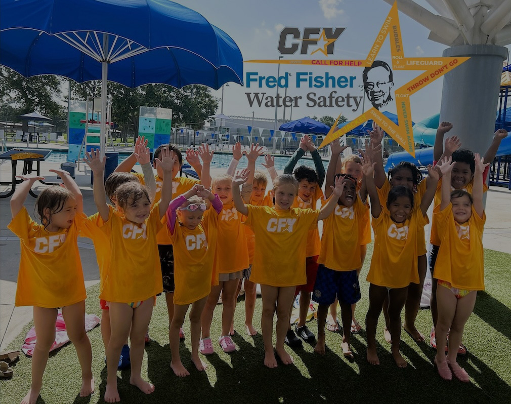 Kids raising their hands up and wearing orange CFY shirts in front of a Fred Fisher Water Safety sign and an outdoor pool.